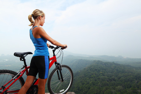 Young sporty lady standing with bicycle on top of a hill and enjoying valley viewの写真素材