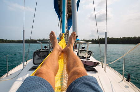 Man relaxing in the hammock set on the sail boat while it anchored near a shoreの写真素材