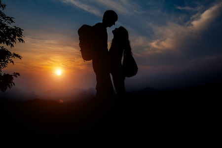 Silhouette of standing couple in love on the mountain with backpacks during sunsetの写真素材