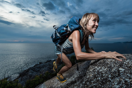 Hiker with backpack climbing natural rocky wall on a dark cloudy background. There are water drops on the skinの写真素材