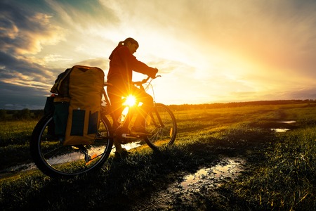 Young lady hiker with loaded bicycle standing on a wet rural road in the meadow at sunsetの写真素材
