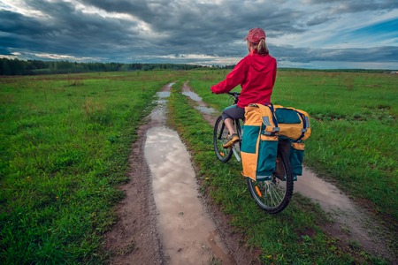 Young lady hiker with loaded bicycle standing on a wet rural road in the meadowの写真素材