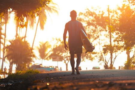 Man with surfboard walks barefoot on the street towards the ocean with trees highlighted by tropical sunの写真素材