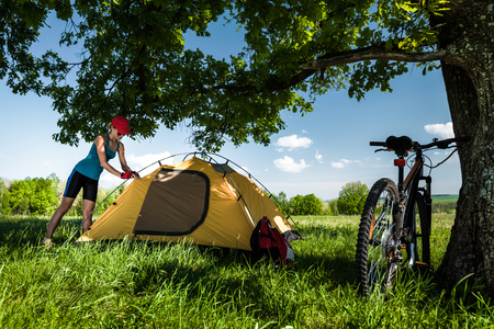 Young lady sets up a tent on a meadow with green grass under treeの写真素材