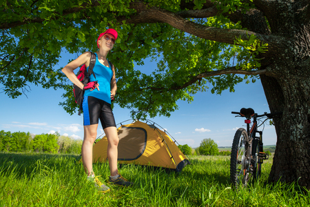 Young lady stands on a meadow with green grass under tree and setted up a tentの写真素材
