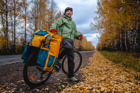 Mature bicycle tourist with his loaded bike on an asphalt autumn roadの写真素材