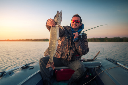 Happy amateur angler holds Pike fish sitting in the boat on the lake during sunriseの写真素材