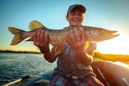 Happy angler holds pike fish sitting in a boat with river on the backgroundの写真素材