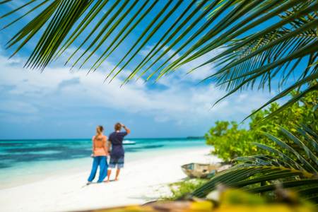 Couple stands on the tropical beach. Focus on the palm tree leaf on the foregroundの写真素材