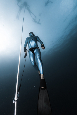 Freediver ascends from a depth along the rope holding a safety leash in a hand. Freedive instructor prepares freedive spot for session, while studens wait on the sea surfaceの写真素材
