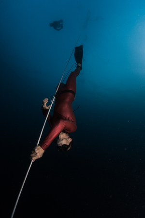 Freediver in red wetsuit descends along the rope. Safety freediver follows him on the background.の写真素材