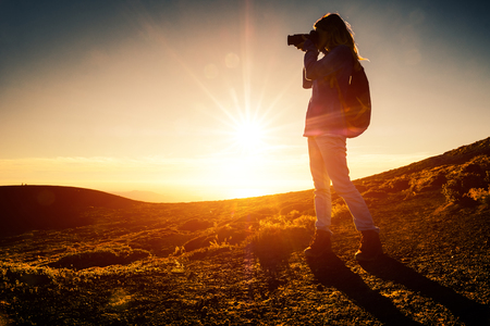 Woman hiker with backpack takes picture of the landscape in mountains during golden hourの写真素材