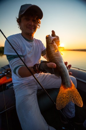 Young angler holds the fish (Zander) sitting in a boat at a lake during sunsetの写真素材