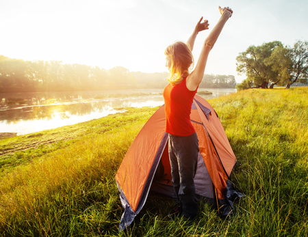 Young woman hiker wakes up and stretches near the tent set on a coast of a riverの写真素材