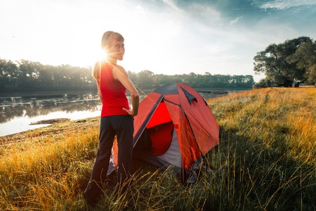 Woman hiker stands near the tent set on a coast of a river and looks at cameraの写真素材