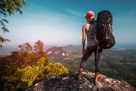 Woman hiker with big backpack stands on the rock and enjoys the valley viewの写真素材