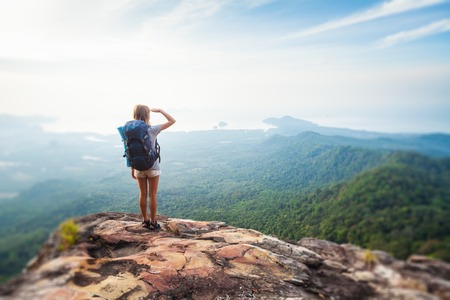 Young woman hiker stands with backpack on the cliff and looks into valley and sea coastline. Tilt shift effect appliedの写真素材
