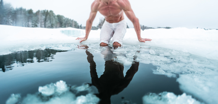 Young man with lean muscular body going to swim in the cold winter water with ice floating on the surface and forest on the background. Tilt shift effect applied, focus on the floating ice piecesの写真素材