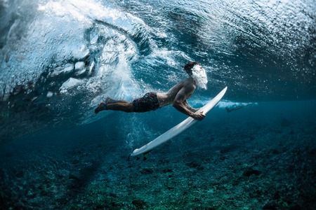 Surfer performs dive (the duck dive) with his surfboard under the wave and exhales air into the water.の写真素材
