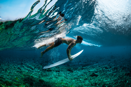 Muscular surfer dives under the crystal clear and glassy ocean wave with his surfboardの写真素材