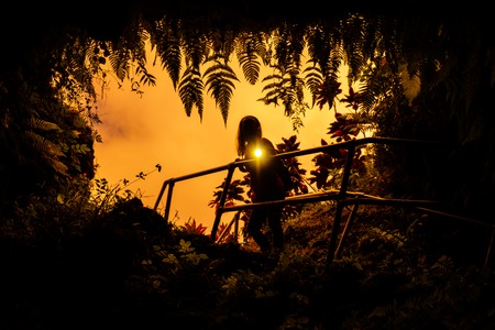 Woman stands near the entrance to the Lava Tube surrounded by lush tropical vegetation and flashes with the light of torch right into the camera. Maui, Hawaiiの写真素材