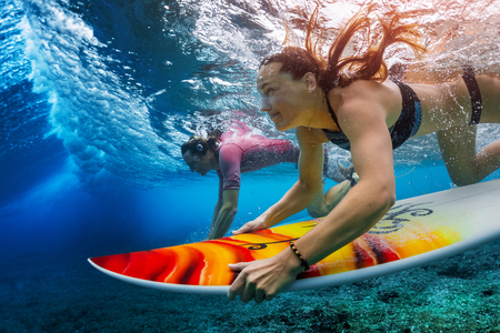 Two young surfers man and woman dive with their surfboards under the breaking ocean waveの写真素材