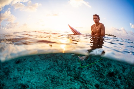 Surfer sits on the surfboard and looks at the camera at sunset. Splitted view with underwater view