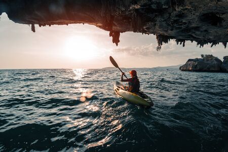 Woman paddles kayak in the tropical sea at sunset and passes the limestone mountains with stalactitesの写真素材