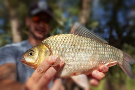 Young happy angler holds the fish (Carassius) and smiles with natural forest backgroundの写真素材