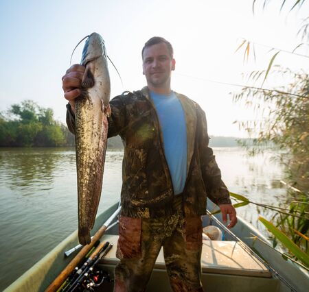 Young amateur angler holds the Wels catfish (Silurus glanis) and look at the camera being in the boat on the river at sunriseの写真素材