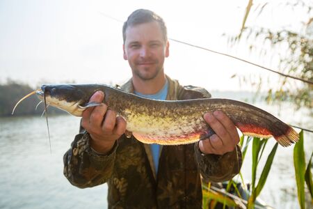 Young amateur angler holds the Wels catfish (Silurus glanis) and look at the camera being in the boat on the river at sunriseの写真素材