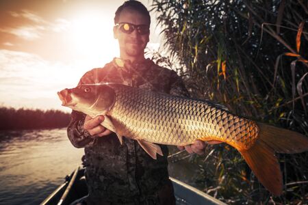 Young amateur angler stands in the boat and holds the big Carp fish (Cyprinus carpio) being on the riverの写真素材