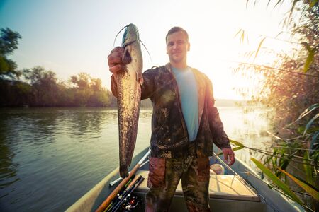 Young amateur angler holds the Wels catfish (Silurus glanis) and look at the camera being in the boat on the river at sunriseの写真素材