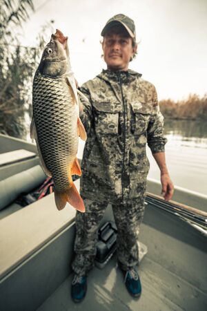 Young amateur angler stands in the boat and holds the big Carp fish (Cyprinus carpio) being on the riverの写真素材