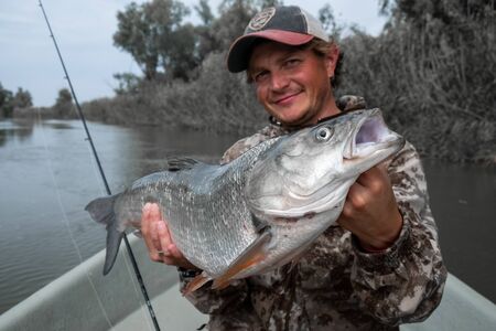 Young angler holds the trophy Asp fish (Leuciscus aspius) being in the boat on the river.の写真素材