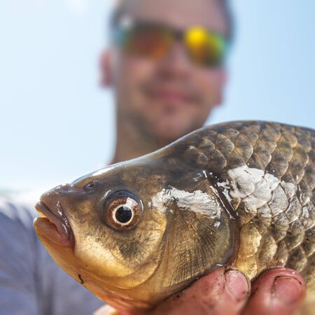 Young happy angler holds the fish (Carassius) and smiles with clear blue sky as the backgroundの写真素材