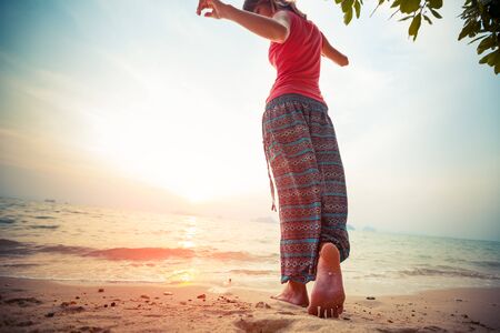 Young woman in hippie pants dances barefoot on the sandy tropical beach at sunset. Focus on the footの写真素材
