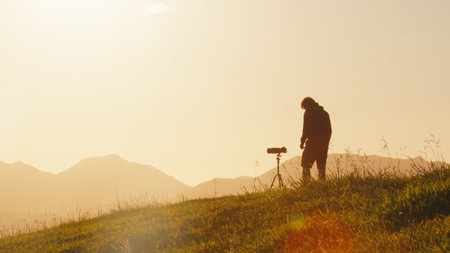 Photographer on the hill. Young male photographer takes pictures with camera and telephoto lens set on tripod on the green hill in the mountains at sunriseの写真素材