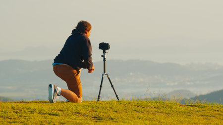 Photographer on the hill. Young male photographer takes pictures with camera and telephoto lens set on tripod on the green hill in the mountains at sunriseの写真素材