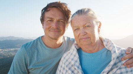 Portrait of young man and elderly woman. Young man hugs senior caucasian woman outdoor at sunrise. Models look into the cameraの写真素材