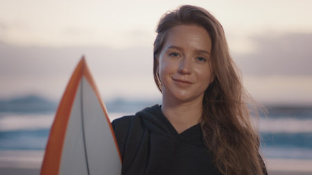 Woman surfer stands with surfing board on the tropical beach in poncho and looks into the cameraの写真素材