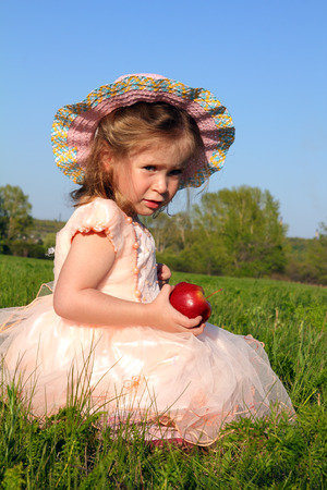 little girl in hat eating apple on meadowの写真素材