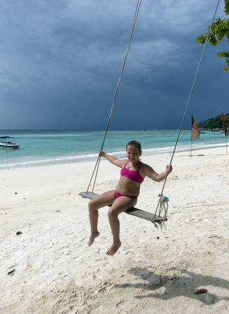 Happy little girl on a swing at the beach with white sand on a background of dark skyの写真素材