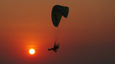 Motor paraglider flying through colorful sunset sky and setting orange sun