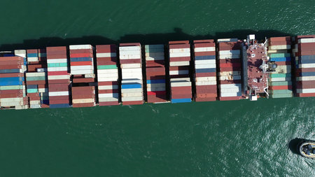 Aerial top view of large cargo ship filled with colorful containers in port area. Small tugboats...