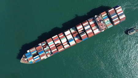 Aerial top view of large cargo ship filled with colorful containers in port area. Small tugboats...