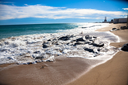 Sunny beach at the day with wet rocks, white clouds and small buildings in the backgroundの写真素材