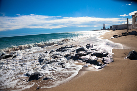 Sunny beach at the day with wet rocks, white clouds and small buildings in the backgroundの写真素材