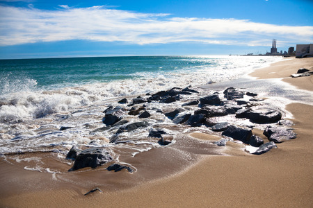 Sunny beach at the day with wet rocks, white clouds and small buildings in the backgroundの写真素材