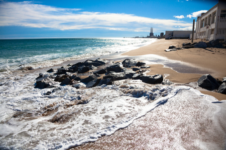 Sunny beach at the day with wet rocks, white clouds and small buildings in the backgroundの写真素材